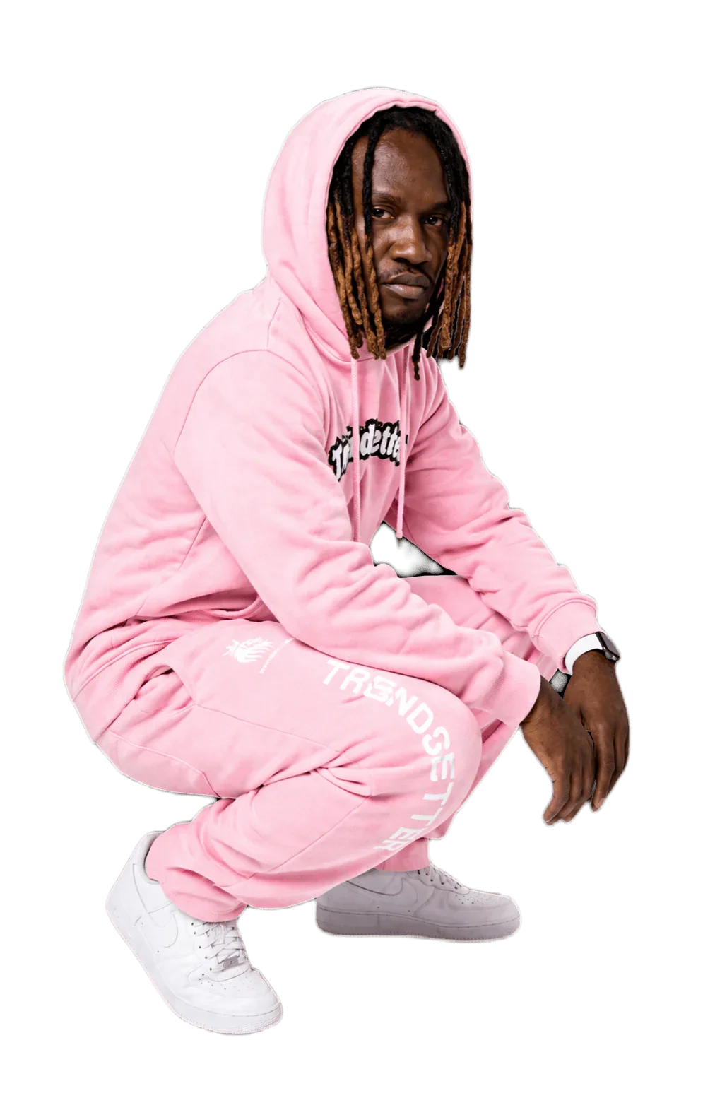 Man in pink matching sweatsuit with 'TRENDSETTER' print, crouching pose, white sneakers, streetwear style